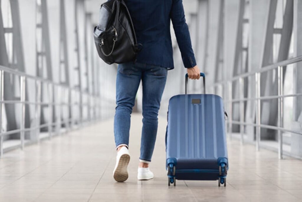 A imagem mostra uma pessoa vista de costas, caminhando por um corredor de aeroporto, que parece ser uma ponte de embarque ou um longo corredor de ligação entre terminais. A pessoa está vestida com um blazer azul escuro sobre uma camisa (não visível), calças jeans azuis e tênis brancos. Ela carrega uma mochila preta de couro ou material similar no ombro esquerdo.
Com a mão direita, a pessoa puxa uma mala de viagem de rodinhas (tipo spinner, com quatro rodas). A mala é de tamanho médio para grande, de cor azul sólida, com uma estrutura rígida e linhas verticais em relevo como detalhe de design. A alça telescópica da mala está estendida.
O ambiente é caracterizado por uma arquitetura moderna, com um piso claro, provavelmente de cerâmica ou material similar, e paredes que são predominantemente estruturas metálicas e vidro, formando grandes janelas ou painéis que se repetem ao longo do corredor, criando uma sensação de profundidade. A iluminação é clara e difusa. A perspectiva da foto é baixa, focando na pessoa e na mala enquanto se movem pelo corredor. Viajar é uma experiência comum, mas é preciso estar ciente de que a Mala sofre danos em Aeroportos devido ao manuseio nos sistemas de bagagem. A imagem captura um momento típico de trânsito em um aeroporto, onde o passageiro se desloca com seus pertences, sempre na esperança de que, ao chegar ao destino, não descubra que sua mala sofreu danos em Aeroportos.