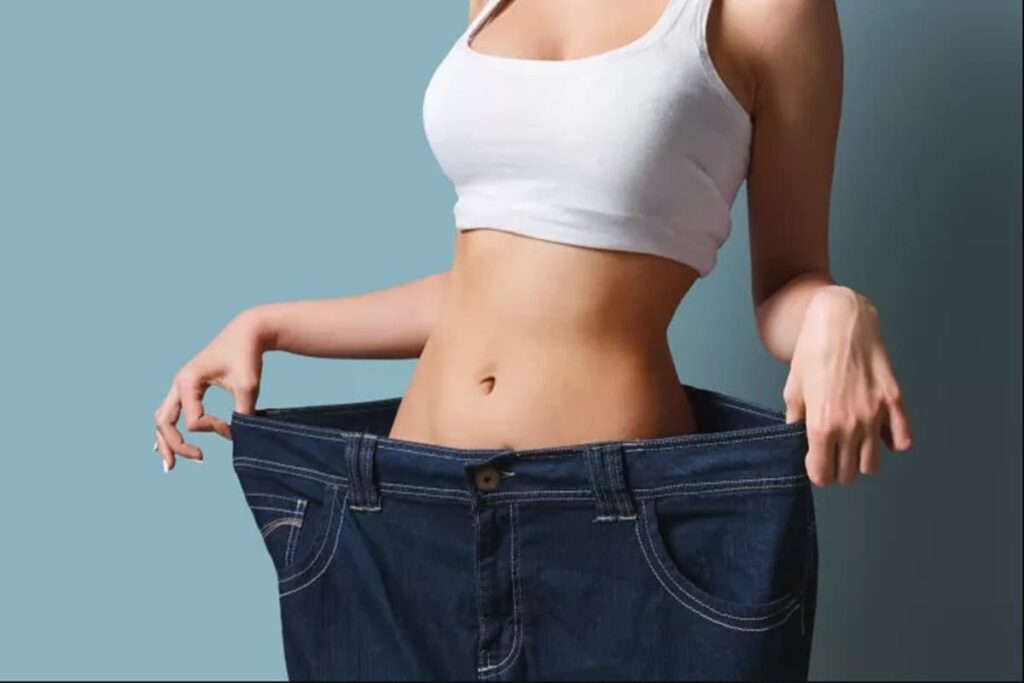 A imagem exibe o tronco de uma mulher com uma silhueta esbelta, posicionada contra um fundo liso de cor azul-acinzentado claro. Ela está vestindo um top branco, estilo regata curta (cropped tank top), que deixa sua barriga à mostra. A parte central da imagem foca em sua cintura fina e abdômen tonificado, com o umbigo visível.
A mulher está dentro de um par de calças jeans de lavagem azul escura. O ponto principal da imagem é que essas calças são dramaticamente grandes para o tamanho atual dela. Com as duas mãos, ela segura as laterais do cós da calça, puxando-o para fora e para os lados, longe de seu corpo. Isso cria um espaço visível e considerável entre a cintura dela e a cintura da calça, enfatizando o quão largas as calças se tornaram.
Detalhes das calças jeans são perceptíveis, como os passadores de cinto, a costura em cor contrastante (possivelmente laranja ou amarela), os bolsos frontais e o botão central acima do zíper. A textura do denim é visível. As mãos da mulher têm unhas bem cuidadas.
A iluminação parece controlada, possivelmente de estúdio, realçando as formas do corpo da mulher e os detalhes da roupa. A composição geral simboliza claramente uma perda de peso significativa, onde as roupas antigas não servem mais. A necessidade de Ajustar Roupas Largas torna-se evidente como resultado dessa transformação física. A imagem transmite uma sensação de conquista, saúde e a consequência prática da mudança corporal.
