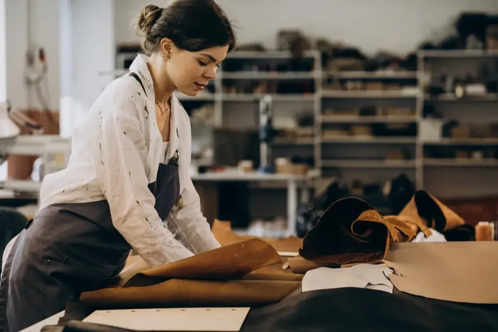 A imagem retrata uma jovem artesã em seu ateliê, imersa em seu trabalho com couro. Ela possui cabelos escuros presos em um coque despojado, com algumas mechas soltas emoldurando seu rosto concentrado. Seu olhar está fixo na peça de couro marrom-claro que ela está manuseando sobre uma grande bancada de trabalho.
Ela veste uma camisa branca de mangas compridas, visivelmente manchada com o que parecem ser respingos de tinta ou corante, um sinal de seu trabalho manual. Por cima da camisa, usa um avental de cor escura, provavelmente azul marinho ou cinza, para proteger suas roupas. Um colar delicado é visível em seu pescoço.
Sobre a bancada, há vários rolos e pedaços de couro de diferentes tonalidades e texturas. Um grande rolo de couro marrom-claro está sendo desenrolado pela artesã. Ao lado, um rolo de couro marrom mais escuro, possivelmente camurça, está enrolado de forma compacta. Pedaços de couro preto e o que parece ser papel pardo para moldes também estão espalhados pela mesa.
O fundo da imagem está levemente desfocado, mas revela um ambiente de oficina bem abastecido. Prateleiras altas e metálicas estão repletas de materiais, ferramentas e possivelmente produtos acabados, indicando um espaço de produção artesanal movimentado e criativo. A iluminação parece ser uma combinação de luz natural e artificial, destacando a figura da mulher e os materiais com os quais ela trabalha. A atmosfera geral é de foco, habilidade e dedicação ao ofício, um local onde a criação e o conserto de roupas de couro em Goiânia poderiam ser habilmente executados.